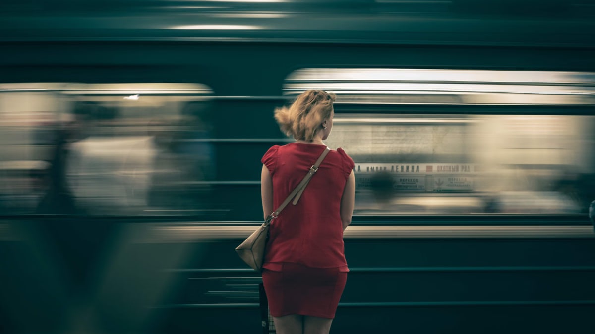 A woman in a red dress waits as a subway train speeds by in a blur, creating a sense of motion.